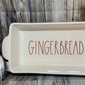 May include: White ceramic loaf pan with red lettering that says "Gingerbread". A red and white gnome with a white beard is to the left of the pan.