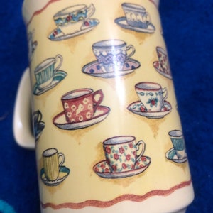May include: A white ceramic mug with a yellow background and a pattern of teacups and saucers. The teacups are decorated with floral and geometric designs in shades of blue, red, green, and yellow.