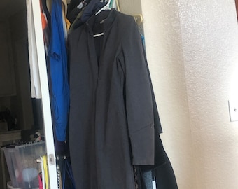 May include: A dark gray long-sleeved jacket hangs on a rack with other clothing. The jacket has a high collar and a zipper closure.
