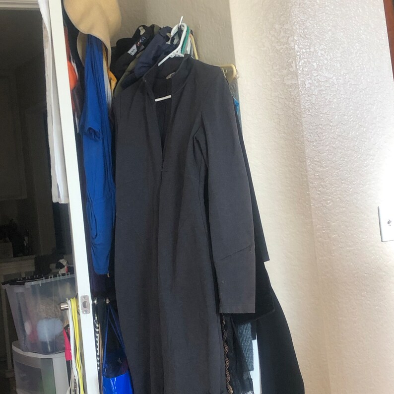 May include: A dark gray long-sleeved jacket hangs on a rack with other clothing. The jacket has a high collar and a zipper closure.