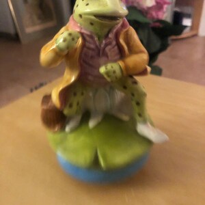 May include: A green frog figurine wearing a yellow jacket and pink vest, sitting on a green lily pad with a blue base.