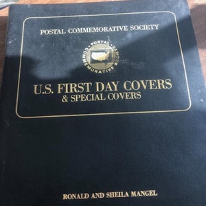 May include: A black album with gold lettering and a gold border. The text reads "POSTAL COMMEMORATIVE SOCIETY" at the top, with "U.S. FIRST DAY COVERS & SPECIAL COVERS" below. The album also features the names "RONALD AND SHEILA MANGEL".