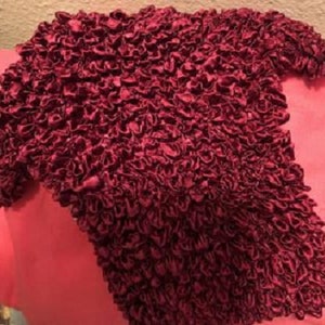 May include: A burgundy-colored, textured, short-sleeved shirt made of a crinkled fabric.