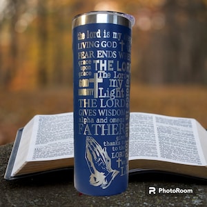 May include: A blue stainless steel tumbler with a silver inscription of a Christian prayer. The tumbler is sitting on top of an open Bible.