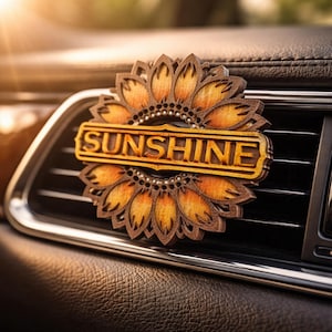 May include: A wooden sunflower car air vent clip with the word "SUNSHINE" in a rectangular frame. The sunflower has a detailed design with brown and yellow petals. The clip is attached to a car's air vent.