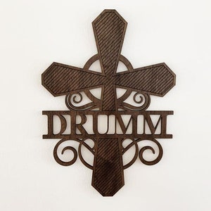 May include: A wooden cross with a decorative scroll design and the name "DRUMM" centered below. The cross is a dark brown color and has a textured finish.