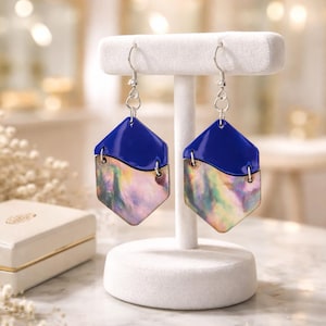 May include: A pair of hexagon-shaped earrings with a two-tone design. The top portion is a solid, glossy blue, while the bottom half features an iridescent, pearlescent finish. The earrings hang from silver-colored hooks and are displayed on a white jewelry stand.