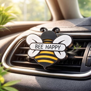 May include: A black, yellow, and white bee-shaped car air vent clip. The clip has a banner that reads "BEE HAPPY". The bee has white wings and a yellow and black striped body. The clip is attached to a car's air vent.