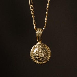 May include: A gold-colored necklace with a round pendant. The pendant has a detailed floral design and a beaded border. The chain is a delicate link style. The background is a dark, blurred setting.