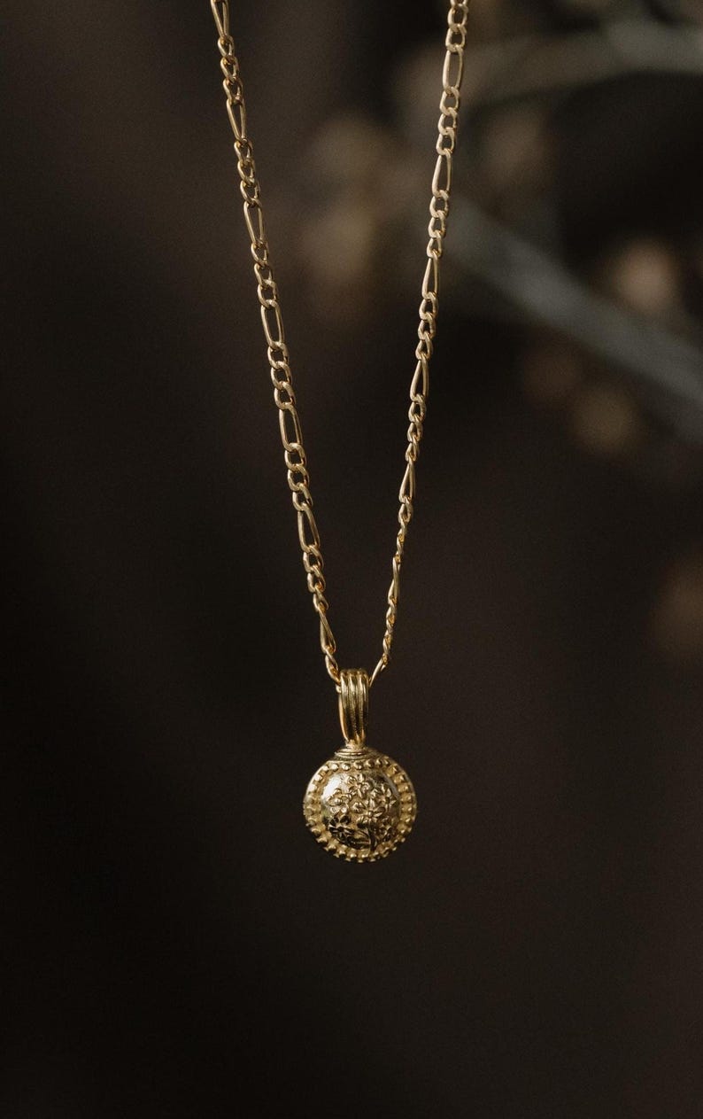 May include: A gold-colored necklace with a round pendant. The pendant has a detailed floral design and a beaded border. The chain is a delicate link style. The background is a dark, blurred setting.
