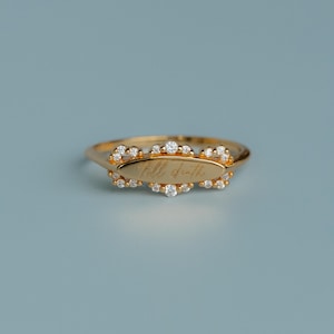 May include: A gold ring featuring an oval plaque with cursive text, surrounded by small, clear gemstones. The ring band is gold, set against a plain, light blue backdrop.
