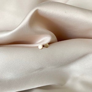 May include: A pair of small, round, gold stud earrings on a white fabric background.