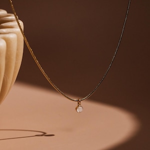 Könnte beinhalten: Eine zarte goldene Kettenkette mit einem kleinen, runden, weißen Edelstein-Anhänger.