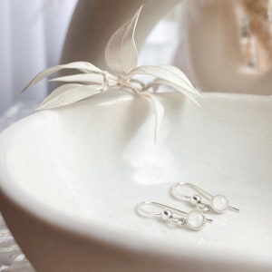 May include: A pair of silver dangle earrings with small, round, white stones. The earrings are displayed on a white ceramic dish.
