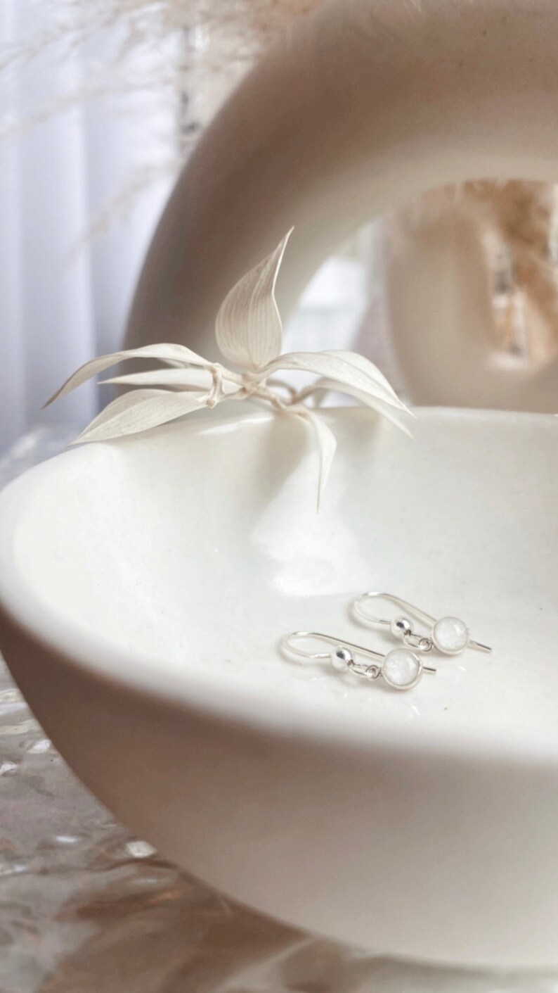 May include: A pair of silver dangle earrings with small, round, white stones. The earrings are displayed on a white ceramic dish.