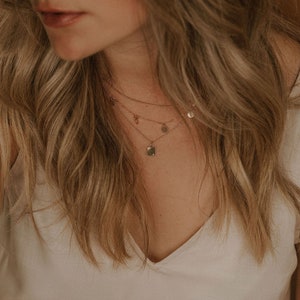 Op de afbeelding: Een vrouw draagt een wit V-hals shirt met een gelaagde gouden ketting met een vierkante hanger en kleine ronde bedels.