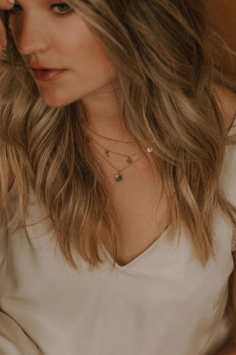 Op de afbeelding: Een vrouw draagt een wit V-hals shirt met een gelaagde gouden ketting met een vierkante hanger en kleine ronde bedels.