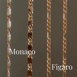 May include: Close-up of three gold-colored chain necklaces. The chains are labeled "Monaco" and "Figaro". The Monaco chain has a unique, textured design, while the Figaro chain has an alternating pattern of long and short links. The chains are displayed against a neutral background.