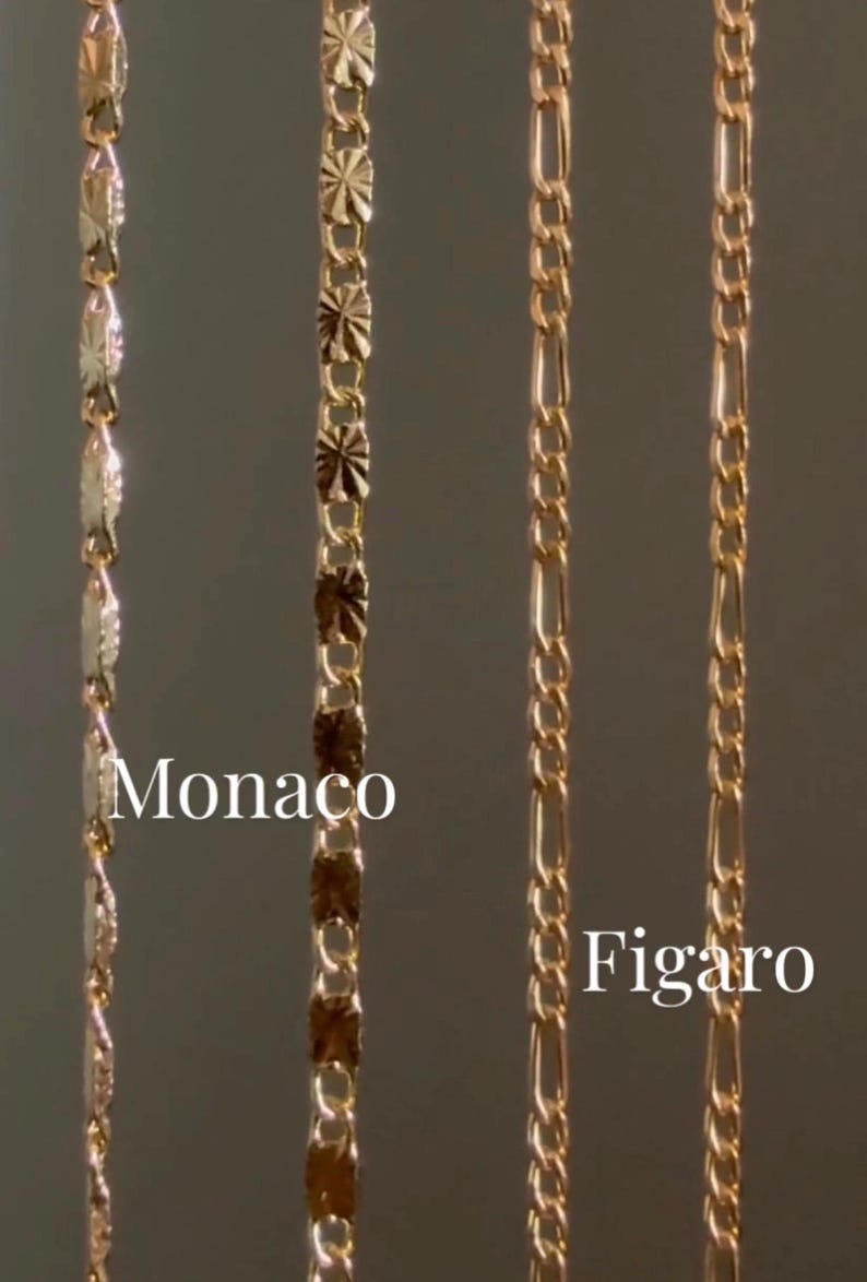 May include: Close-up of three gold-colored chain necklaces. The chains are labeled "Monaco" and "Figaro". The Monaco chain has a unique, textured design, while the Figaro chain has an alternating pattern of long and short links. The chains are displayed against a neutral background.