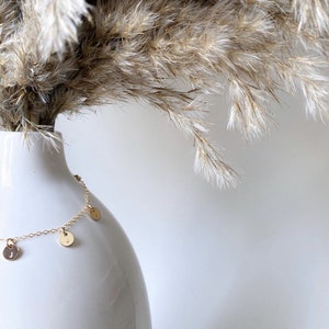 Op de afbeelding: Een gouden ketting met kleine ronde bedels die eraan hangen. De bedels hebben letters erop. De ketting wordt getoond op een witte vaas met gedroogd pampasgras.