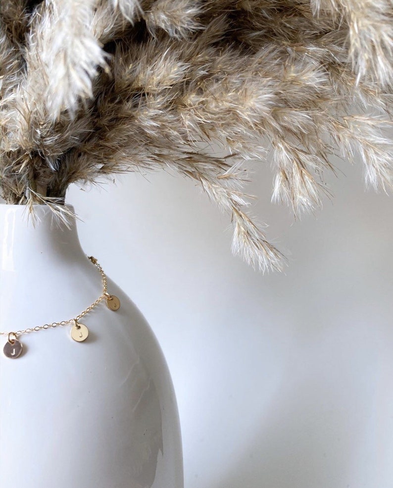Op de afbeelding: Een gouden ketting met kleine ronde bedels die eraan hangen. De bedels hebben letters erop. De ketting wordt getoond op een witte vaas met gedroogd pampasgras.