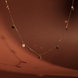 Op de afbeelding: Een delicate gouden ketting met kleine, ronde, gouden bedels die eraan hangen. De ketting ligt op een bruine achtergrond.
