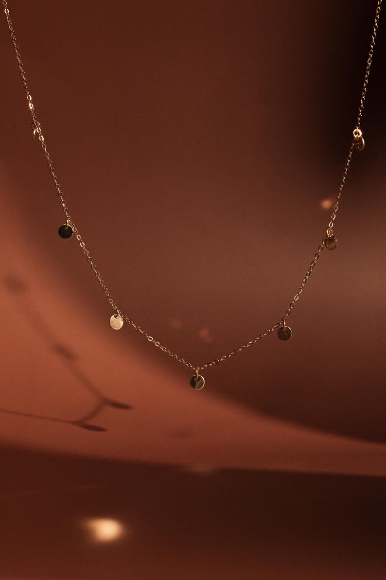 Op de afbeelding: Een delicate gouden ketting met kleine, ronde, gouden bedels die eraan hangen. De ketting ligt op een bruine achtergrond.
