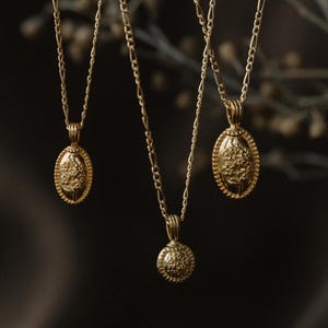 May include: Three gold-colored necklaces with different pendant designs. One has a round pendant, and two have oval pendants. Each pendant features a floral design. The necklaces hang against a dark background, highlighting the jewelry's details.