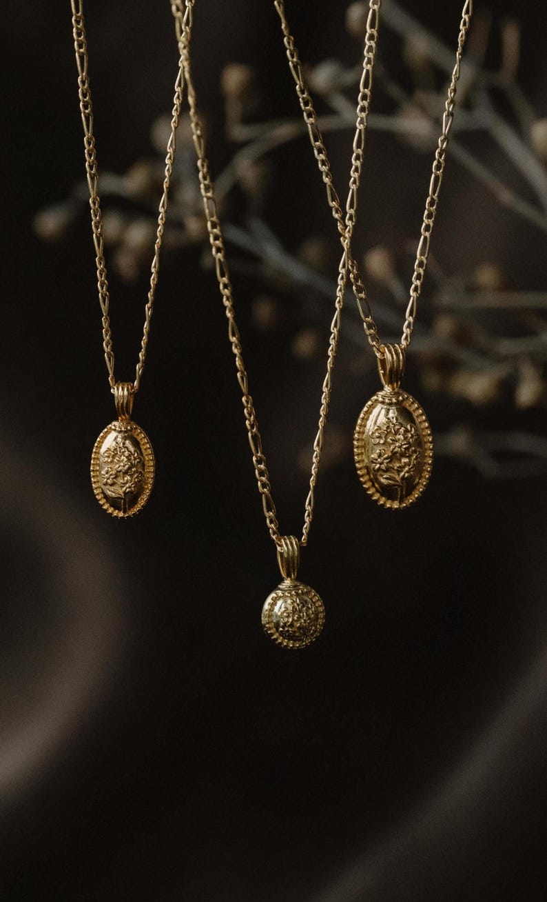 May include: Three gold-colored necklaces with different pendant designs. One has a round pendant, and two have oval pendants. Each pendant features a floral design. The necklaces hang against a dark background, highlighting the jewelry's details.