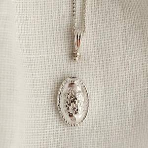 May include: A silver-colored necklace with an oval pendant. The pendant features a detailed floral design within a beaded border. The chain and pendant are displayed against a white, textured background.
