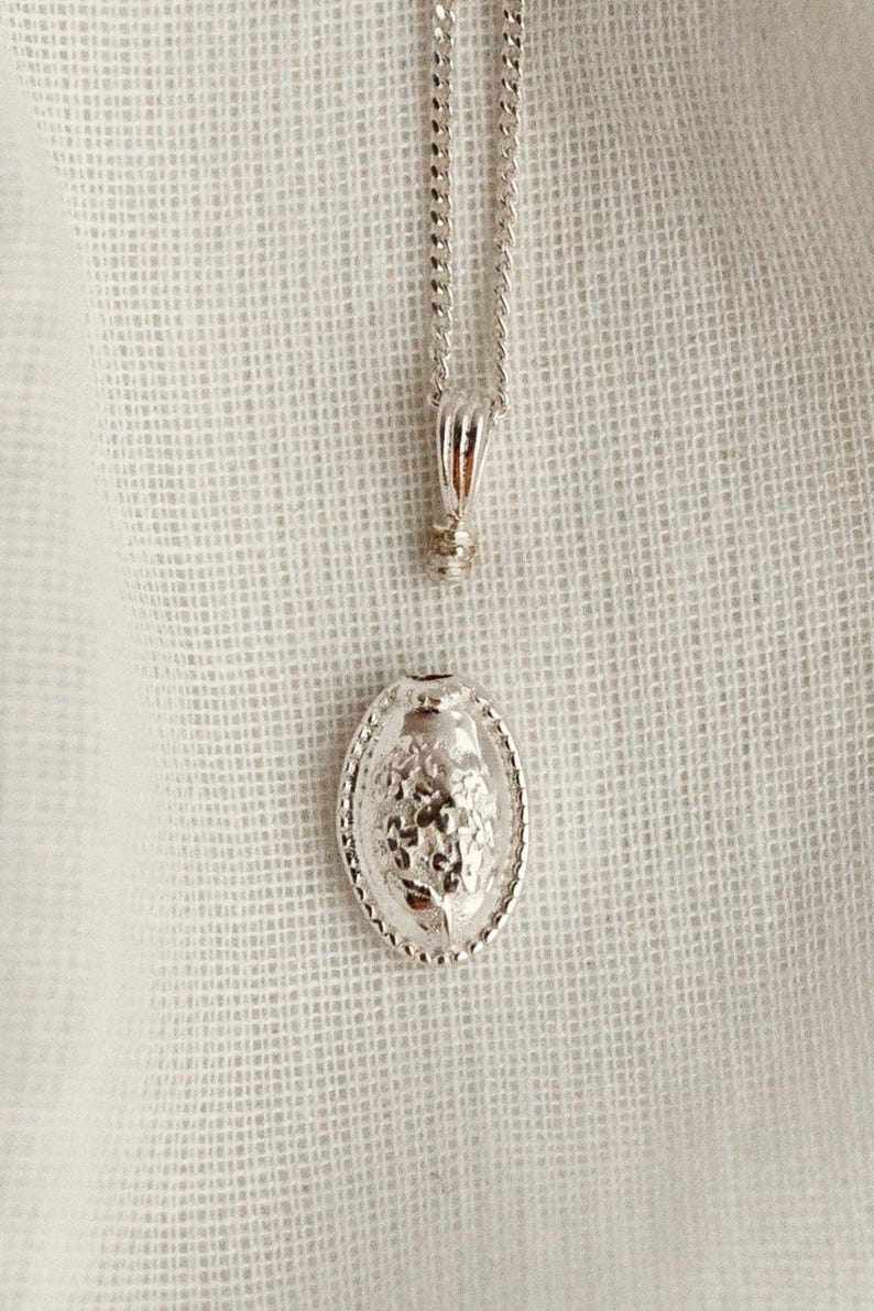 May include: A silver-colored necklace with an oval pendant. The pendant features a detailed floral design within a beaded border. The chain and pendant are displayed against a white, textured background.