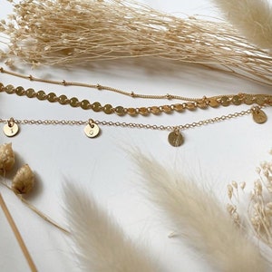 Op de afbeelding: Drie gouden kettingen met delicate kettingen. De bovenste ketting heeft een ketting van kleine gouden cirkels. De twee onderste kettingen hebben elk &eacute;&eacute;n bedel, met de letters 'I' en 'G' erop gegraveerd.