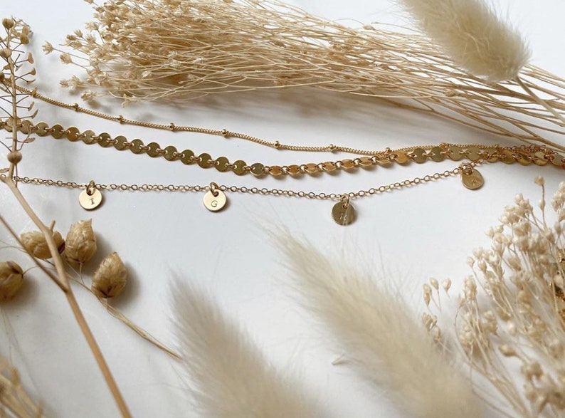 Op de afbeelding: Drie gouden kettingen met delicate kettingen. De bovenste ketting heeft een ketting van kleine gouden cirkels. De twee onderste kettingen hebben elk &eacute;&eacute;n bedel, met de letters 'I' en 'G' erop gegraveerd.