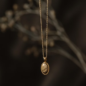 Puede incluir: Un collar dorado con una cadena delicada y un colgante ovalado. El colgante presenta un diseño floral detallado dentro de un borde de cuentas. El collar cuelga sobre un fondo oscuro borroso, resaltando los intrincados detalles de la joya.