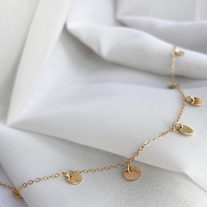 Op de afbeelding: Een delicate gouden ketting met kleine, ronde, gouden bedels die eraan hangen. De ketting ligt op een witte stoffen achtergrond.