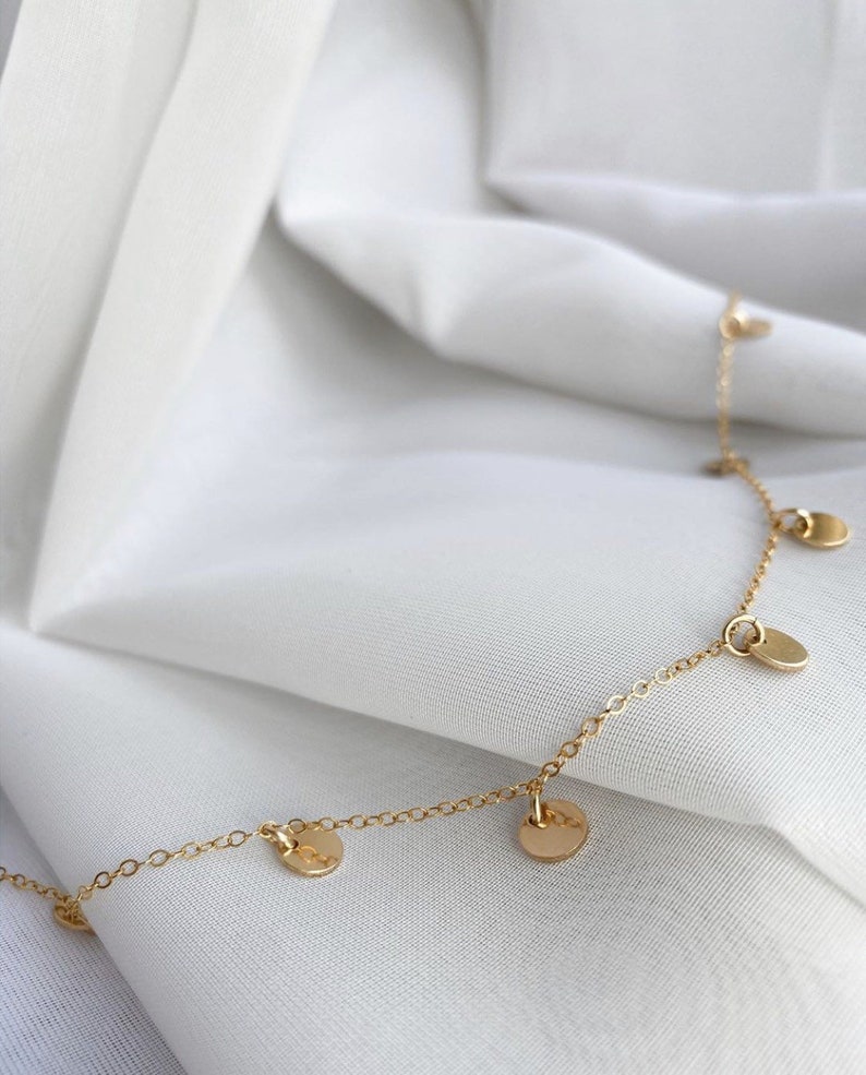 Op de afbeelding: Een delicate gouden ketting met kleine, ronde, gouden bedels die eraan hangen. De ketting ligt op een witte stoffen achtergrond.
