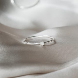 May include: A slender silver ring featuring a small, circular, white gemstone. The ring is displayed on a backdrop of soft, silvery-white fabric, creating a minimalist and elegant aesthetic. The ring's thin band and simple design suggests understated beauty.