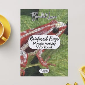 Könnte beinhalten: Ein Arbeitsbuch mit dem Titel "Rainforest Frogs Mosaic Activity Workbook" mit einem Froschmosaik-Design auf dem Cover. Das Cover zeigt einen rot-weißen Frosch auf einem grünen Mosaikhintergrund. Eine gelbe Tasse und Untertasse befinden sich links.