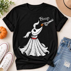 May include: A black t-shirt featuring a white graphic of a ghost dog with a red collar. A skeletal hand reaches out to touch the dog's nose, which is a small orange pumpkin. The word Boop is printed in white script above the dog's head.