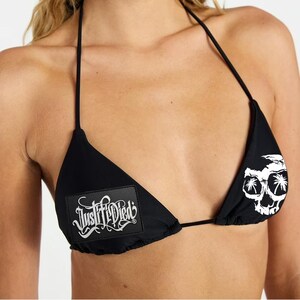 May include: Black bikini top with a skull and palm tree graphic on one cup and the text "Justified Died" on the other. The bikini top has thin black straps.
