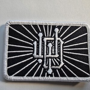 May include: Black and white embroidered patch with a rectangular shape. The design features a stylized emblem in white, set against a black background with radiating white lines. The patch has a white border.