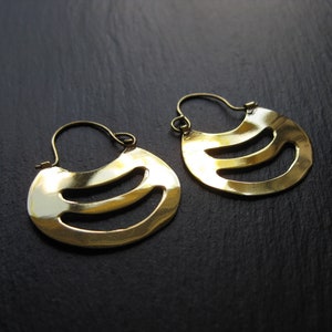 May include: A pair of gold hoop earrings with a unique design featuring two curved lines cut out of the metal. The earrings are on a black background.