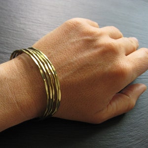 Peut inclure: Un ensemble de cinq bracelets en métal doré superposés au poignet d'une personne.