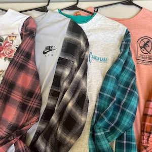 May include: Four long-sleeve shirts hanging on black hangers. The first is white with a floral print and red plaid sleeves. The second is light gray with a Nike Air logo and black plaid sleeves. The third is heather gray with Crater Lake text and teal plaid sleeves. The fourth is peach with a Route of the Hiawatha graphic and orange plaid sleeves.