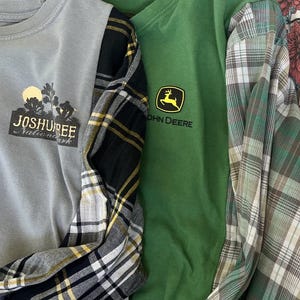 May include: A gray long-sleeve shirt with a Joshua Tree National Park graphic and black and yellow plaid sleeves, next to a green John Deere t-shirt with a yellow deer logo and green and white plaid sleeves, both on black hangers.
