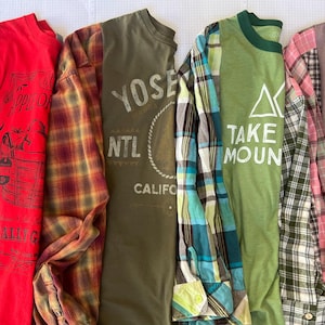 May include: Four long-sleeved shirts with mixed patterns and solid colors. The first is red with a black plaid sleeve and apple basket graphic. The second is olive green with a plaid sleeve and Yosemite text. The third is green with a plaid sleeve and mountain graphic. The fourth is mauve with a pink plaid sleeve and floral text.