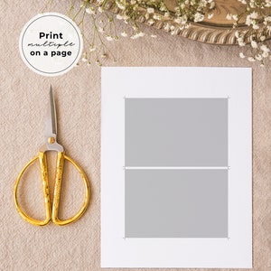 May include: A white sheet of paper with two gray rectangles, each with a thin white line separating them. The paper is on a beige surface with a pair of gold scissors and a white flower arrangement in the background.