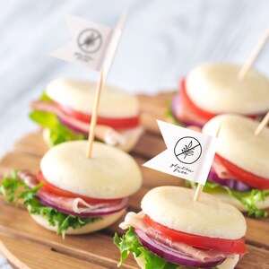 May include: Close-up of four mini mozzarella cheese sandwiches on a wooden board. Each sandwich is topped with lettuce, tomato, red onion, and prosciutto. Two of the sandwiches have a small white flag with the words "gluten free" written on them.