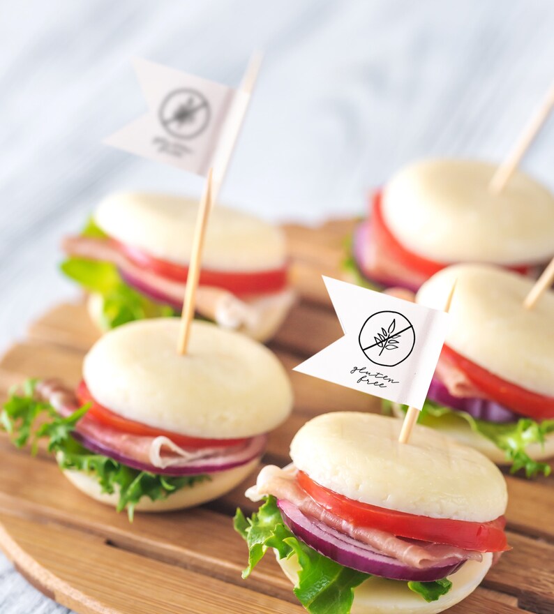 May include: Close-up of four mini mozzarella cheese sandwiches on a wooden board. Each sandwich is topped with lettuce, tomato, red onion, and prosciutto. Two of the sandwiches have a small white flag with the words "gluten free" written on them.