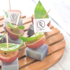 May include: A wooden platter with small purple and white squares topped with red grapes, basil leaves, and toothpicks with small flags that say "vegan".